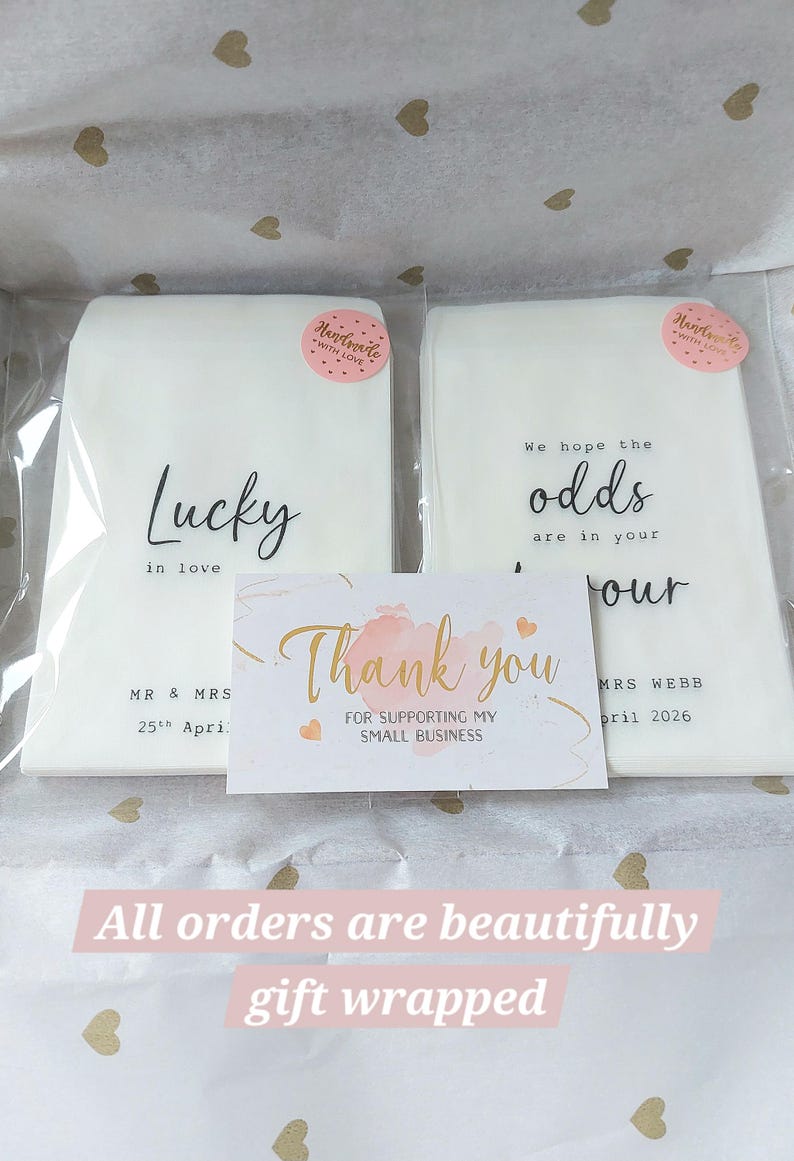 May include: Two white rectangular paper packets in clear plastic sleeves, each with a pink 'Handmade with love' sticker. The left packet reads 'Lucky in love MR & MRS 25th April'. The right reads 'We hope the odds are in your favour MRS WEBB April 2026'. A 'Thank you' card is centered in front.