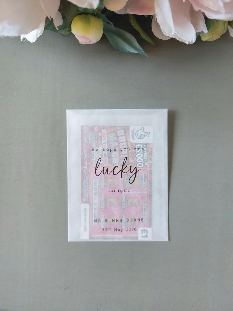 May include: A rectangular white translucent paper envelope containing a pink lottery scratch card. The front of the envelope features black printed text that reads: We hope you get lucky tonight, MR & MRS DEANE, 30th May 2026.