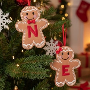 May include: Two tan felt gingerbread person ornaments hanging from a Christmas tree. One ornament features a red letter N and a red bow tie, while the other features a red letter E and a red hair bow. Both have white stitched icing details.