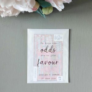 May include: A rectangular white paper envelope containing a lottery scratch card. The front of the envelope features the printed text "We hope the odds are in your favour" followed by "AMELIA & JAMES" and the date "3rd June 2026" in black ink.