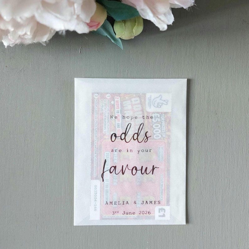 May include: A rectangular white paper envelope containing a lottery scratch card. The front of the envelope features the printed text "We hope the odds are in your favour" followed by "AMELIA & JAMES" and the date "3rd June 2026" in black ink.