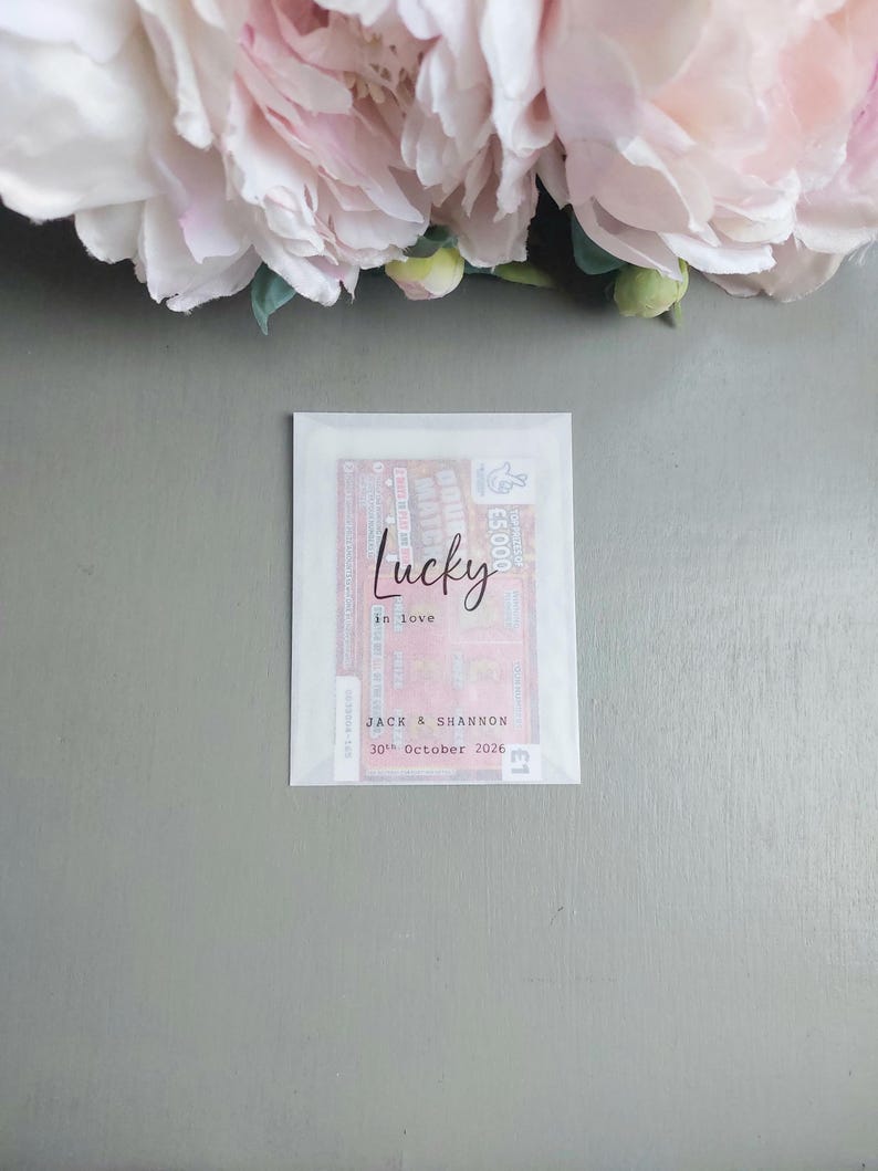 May include: A rectangular translucent paper envelope containing a lottery ticket. The front of the envelope features the black printed text Lucky in love, followed by the names Jack & Shannon and the date 30th October 2026 in a typewriter font.