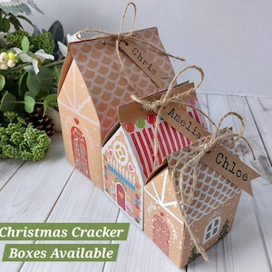 May include: Three house-shaped cardboard gift boxes with gingerbread-inspired designs, featuring red and white accents, scalloped roof patterns, and brown paper tags tied with twine labeled with the names Chris, Amelia, and Chloe.