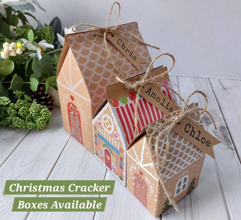 May include: Three house-shaped cardboard gift boxes with gingerbread-inspired designs, featuring red and white accents, scalloped roof patterns, and brown paper tags tied with twine labeled with the names Chris, Amelia, and Chloe.