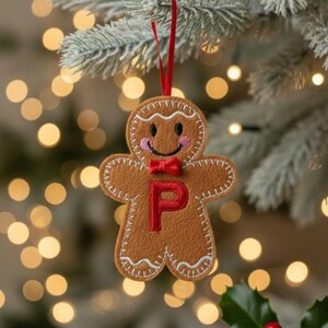 May include: A brown felt gingerbread man ornament featuring white stitched detailing, pink embroidered cheeks, a small red fabric bow tie, and a prominent red letter P stitched in the center. The ornament hangs from a thin red ribbon loop.