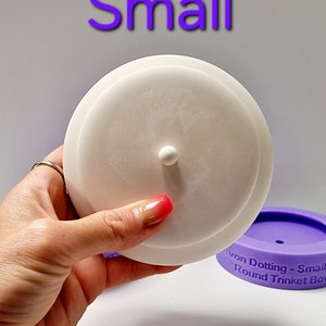 Może przedstawiać: Biała silikonowa pokrywka do małej, okrągłej miseczki na drobiazgi. Pokrywka ma mały guzik na górze. Miseczka jest fioletowa i ma napis "Avon Dotting - Small Round Trinket Bowl" na boku.