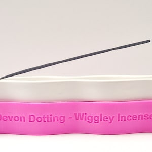 Puede incluir: Porta incienso de cerámica blanca con una base de silicona rosa. La base tiene el texto "Devon Dotting - Wigglely Incense" impreso en ella. Un palo de incienso negro está apoyado en el porta incienso.