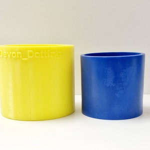 Könnte beinhalten: Zwei zylindrische Behälter, einer gelb und einer blau. Der gelbe Behälter hat den Text "Devon_Dotting" auf der Seite.