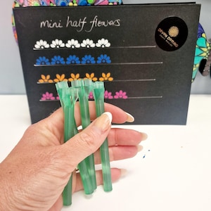 Puede incluir: Una mano sostiene varias herramientas verdes translúcidas frente a una tarjeta negra que muestra las palabras "mini half flowers" y filas de diseños florales coloridos. La tarjeta tiene una pegatina circular con las palabras "DEVON DOTING".
