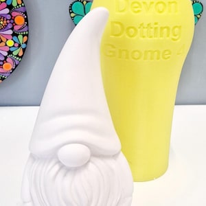 May include: A white ceramic gnome figurine with a long, pointed hat and a bushy beard. The gnome is next to a yellow silicone mold with the text "Devon Dotting Gnome" on it.