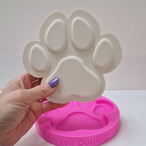 May include: A white ceramic paw print mold being placed into a pink silicone paw print mold. The silicone mold has the text "Devon Dotting" on the bottom.