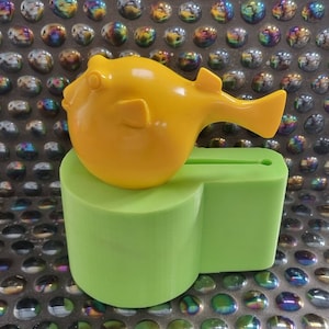 Peut inclure: Une figurine de poisson-globe jaune avec une base verte. La base a une fente pour tenir un téléphone ou un autre appareil.