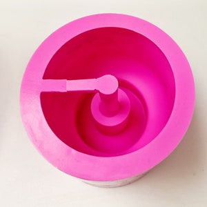 May include: A bright pink, circular silicone mold with a central post and a side extension. The mold has a smooth, matte finish and is designed for crafting or baking. The design features a rounded interior and a sturdy base.