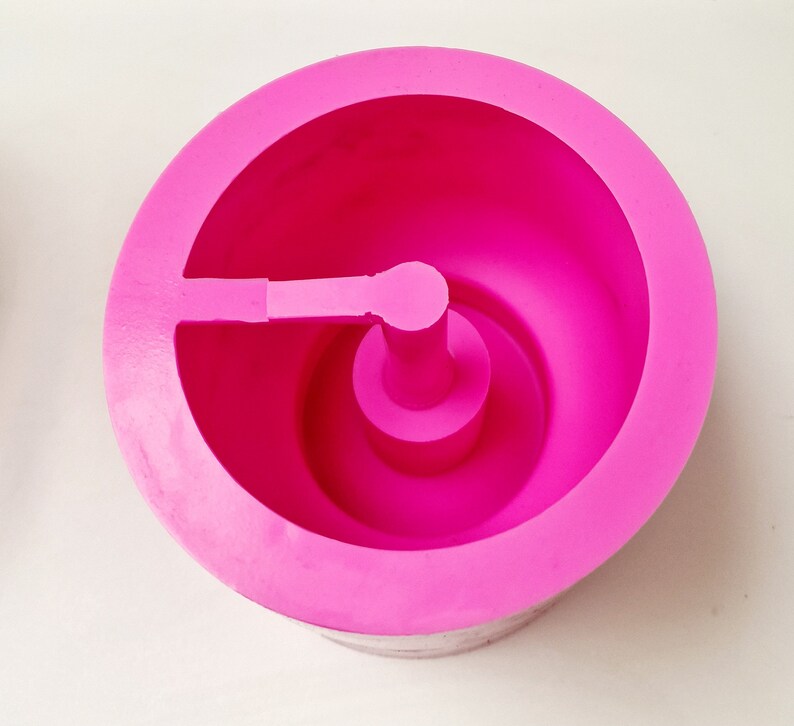 May include: A bright pink, circular silicone mold with a central post and a side extension. The mold has a smooth, matte finish and is designed for crafting or baking. The design features a rounded interior and a sturdy base.
