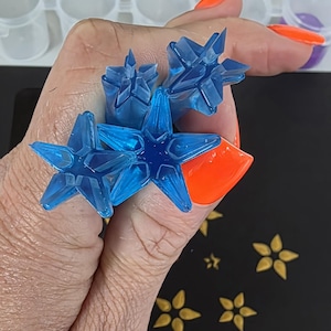 May include: Several translucent blue star-shaped resin pieces are held in a hand with orange painted nails. The stars have a faceted design. Gold star-shaped embellishments are scattered on a black surface.