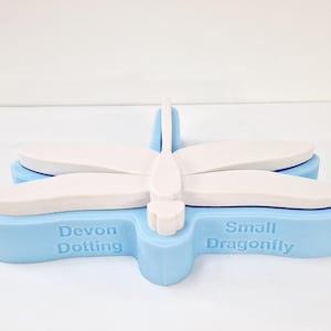 May include: A light blue and white dragonfly-shaped object. The object has a light blue base with the words "Devon Dotting" and "Small Dragonfly" printed on it. The wings and body are white.