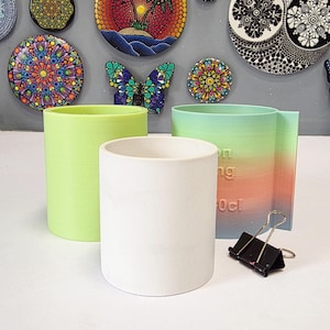 May include: A collection of colorful, decorative wall art and cylindrical planters. The planters are in shades of green, white, and a gradient of orange and green. A black binder clip is also present.
