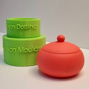 Könnte beinhalten: Zwei grüne Silikonformen, die übereinander gestapelt sind, mit dem Text "Devon Dotting" und "Devon Moulds". Eine leuchtend rote Silikonform in Form einer Kuppel mit einem Knopf oben.