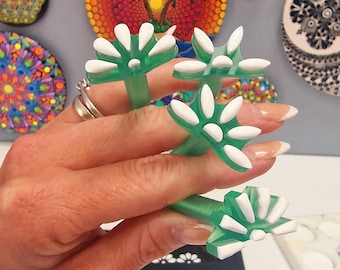 May include: Green and white flower-shaped rings are displayed in the image. The rings are on a hand with long, manicured nails. In the background, there are several decorative, colorful, circular art pieces.