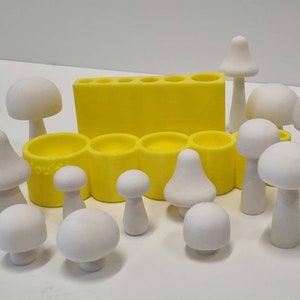 以下が含まれることがあります： 様々なサイズで、一部はベル型の帽子をかぶった白いセラミック製のキノコの置物のセット。黄色いプラスチック製の型に、複数の円形のくぼみが並んでいます。