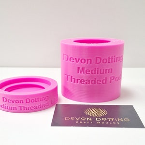 May include: Two pink silicone molds for making candles. The larger mold is a cylinder with the text "Devon Dotting Medium Threaded Pot" on the side. The smaller mold is a ring with the text "Devon Dotting Medium Threaded Lid" on the side.