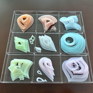 Op de afbeelding: Een doorzichtige acryl display case met negen compartimenten, elk met een unieke, gelaagde abstracte sculptuur. De sculpturen hebben pastelkleuren zoals perzik, roze, groen en blauw, met verschillende geometrische en organische vormen.