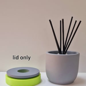 Może przedstawiać: Dyfuzor ceramiczny w kolorze szarym z czarnymi trzcinami i szarą pokrywką. Pokrywka jest oddzielna i pokazana na zielono-szarej podstawie. Na zdjęciu widnieje tekst "lid only".