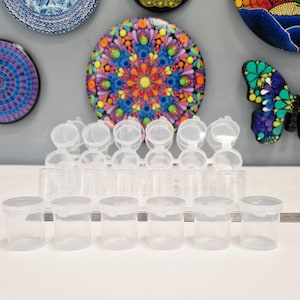 May include: A selection of small, clear plastic containers with hinged lids. The containers are cylindrical and arranged in rows. The lids are white and snap shut. The background features colourful decorative wall art.