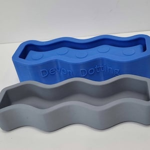 Puede incluir: Dos moldes de silicona, uno azul y otro gris, con forma de línea ondulada. El molde azul tiene cuatro hendiduras circulares y el texto "Devon Dotting" en la parte inferior.