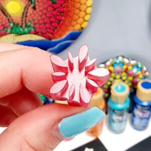 May include: A pink rubber stamp with a flower design. The stamp is being held by a hand with blue nail polish. The stamp is used for creating dot art on rocks.