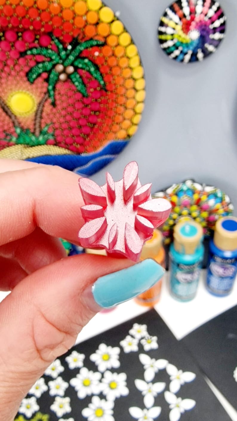 May include: A pink rubber stamp with a flower design. The stamp is being held by a hand with blue nail polish. The stamp is used for creating dot art on rocks.