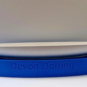 Puede incluir: Un plato de cerámica blanco de forma ovalada se asienta sobre una base azul de forma ovalada. La base tiene las palabras "Devon Dotting" en relieve en la parte delantera.