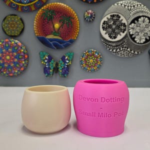 Puede incluir: Dos pequeñas macetas redondas. Una es de color crema blanquecino, y la otra es de color rosa brillante con el texto "Devon Dotting - Small Milo Pot" impreso en el lateral. Arte decorativo de pared en el fondo.
