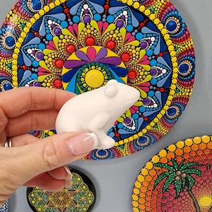 Könnte beinhalten: Eine Sammlung von bunten, runden bemalten Steinen mit Mandala-Mustern. Die Steine sind mit Punkten und Spiralen in verschiedenen Farben verziert, darunter Rot, Gelb, Blau, Grün und Lila. Eine weiße Keramik-Froschfigur wird vor den Steinen gehalten.