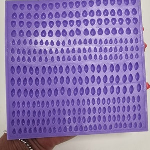 Puede incluir: Molde de silicona morado con múltiples cavidades pequeñas en forma de lágrima. El molde está diseñado para crear pequeños artículos decorativos.