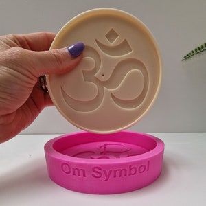 Puede incluir: Un molde de silicona rosa con las palabras "Om Symbol" en el lateral. El molde está diseñado para crear una fundición de resina blanca del símbolo Om.