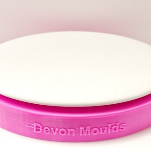 May include: A pink silicone cake mould with a white removable base. The mould has the words "Devon Moulds" printed on the side.