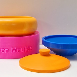 Puede incluir: Un juego de tres moldes de silicona para hacer velas. Los moldes tienen forma de cuenco, cilindro y cono. Los moldes de cuenco y cono son de color naranja, el molde de cilindro es de color rosa y el molde de cono tiene un interior azul. El molde de cilindro tiene las palabras "Devon Moulds" en relieve.