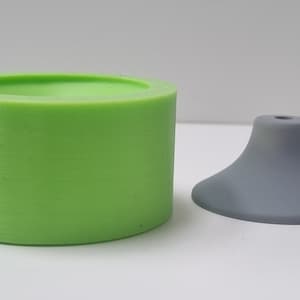 Könnte beinhalten: Eine grüne Silikonform zum Herstellen einer Kerze oder Seife, neben einem grauen Kunststoffsockel für eine Kerze oder Seife.
