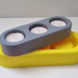May include: A gray candle holder with three candle wells sits on top of a yellow silicone mold. The mold has the text "Devon_Dotting" embossed on the bottom.