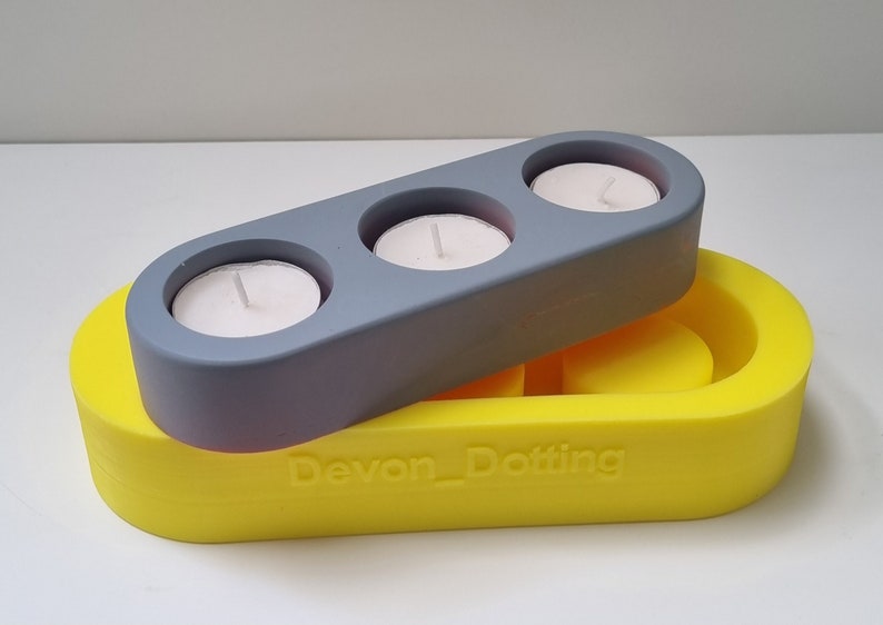 May include: A gray candle holder with three candle wells sits on top of a yellow silicone mold. The mold has the text "Devon_Dotting" embossed on the bottom.