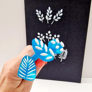 May include: A blue plastic tool with white leaf-shaped stamps. The tool is used to create leaf patterns on a black surface.