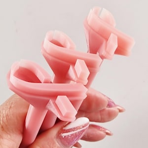 Puede incluir: Tres herramientas rosas y geométricas para nail art se sostienen en una mano. Las herramientas tienen una forma única con una parte superior curva y una base rectangular. El fondo es blanco liso. La mano tiene uñas largas con un diseño rosa y blanco.