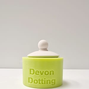 May include: A green cylindrical container with a white lid and a knob on top. The container has the text "Devon Dotting" printed on the side.