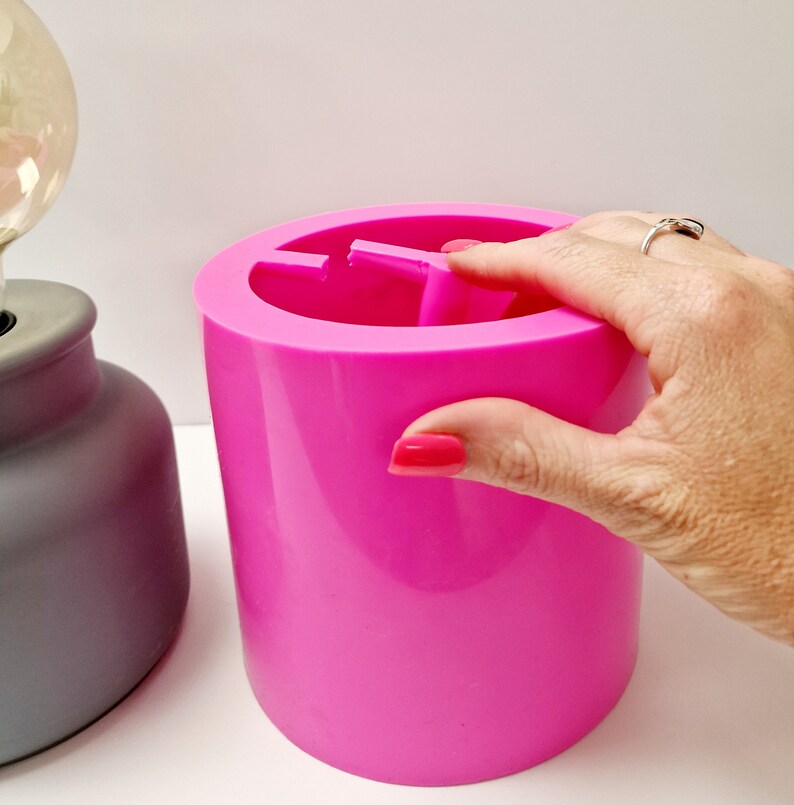 May include: A bright pink, cylindrical container with a divided interior. The container is smooth and solid, with a hand partially visible on the side. A gray lamp base is visible in the background.