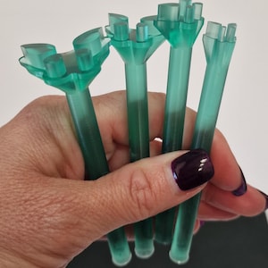 May include: Four translucent teal plastic tools are held in a hand. Each tool has a thin stem and a multi-pronged head. The hand has dark purple nail polish. The tools are likely for a specific craft.