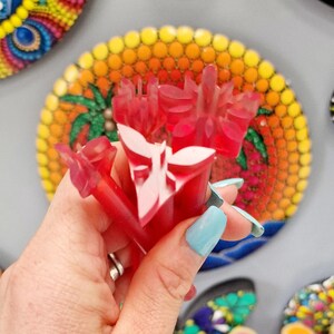 May include: A set of red plastic dotting tools with white designs. The tools are held in a hand with blue nail polish. The tools are in front of a colorful dot painting.