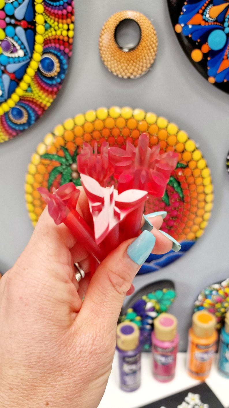 May include: A set of red plastic dotting tools with white designs. The tools are held in a hand with blue nail polish. The tools are in front of a colorful dot painting.