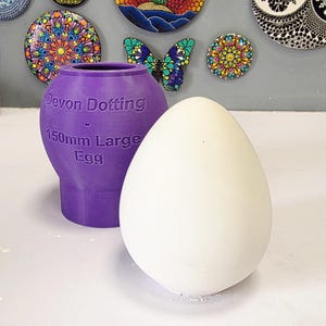 Pode incluir: Um ovo branco grande ao lado de um recipiente roxo com o texto "Devon Dotting - 150mm Large Egg". O recipiente tem formato de vaso. Arte mandala decorativa no fundo.