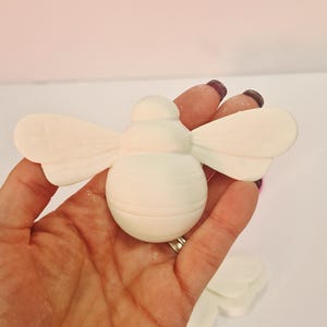 May include: A white, bee-shaped decorative item with detailed wings and body segments. The bee is held in a hand against a light pink background. The item appears to be made of a smooth, solid material.